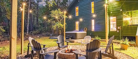 Ein gemütlicher Sitzbereich im Freien mit Adirondack-Stühlen um eine Feuerstelle, einer Holzterrasse und einem Haus im Hintergrund.