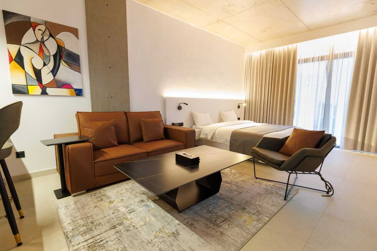 Een moderne hotelkamer met een bruine bank, een klein bijzettafeltje, een bed met witte lakens en een groot raam met gordijnen.