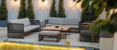 An outdoor seating area with a pool, surrounded by tall plants and wall-mounted lights.