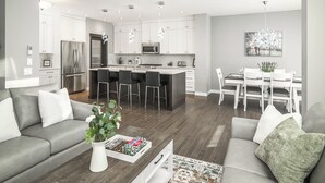 A modern kitchen with stainless steel appliances, a dining area with white chairs, and a gray sofa with decorative pillows.