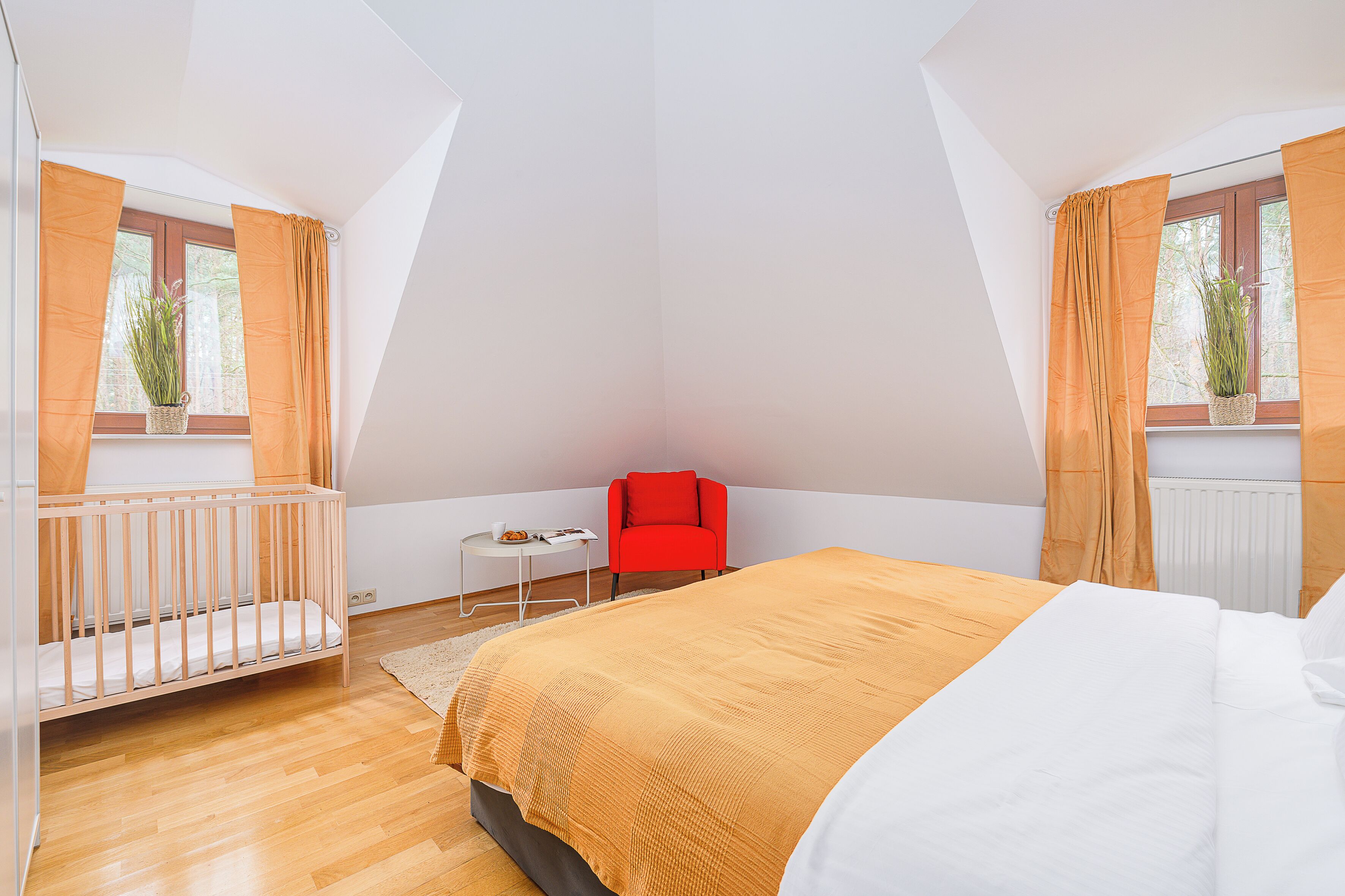 A bedroom with a wooden floor, a bed with a white and orange bedspread, a red chair, a small table with a plant, and a window with curtains.