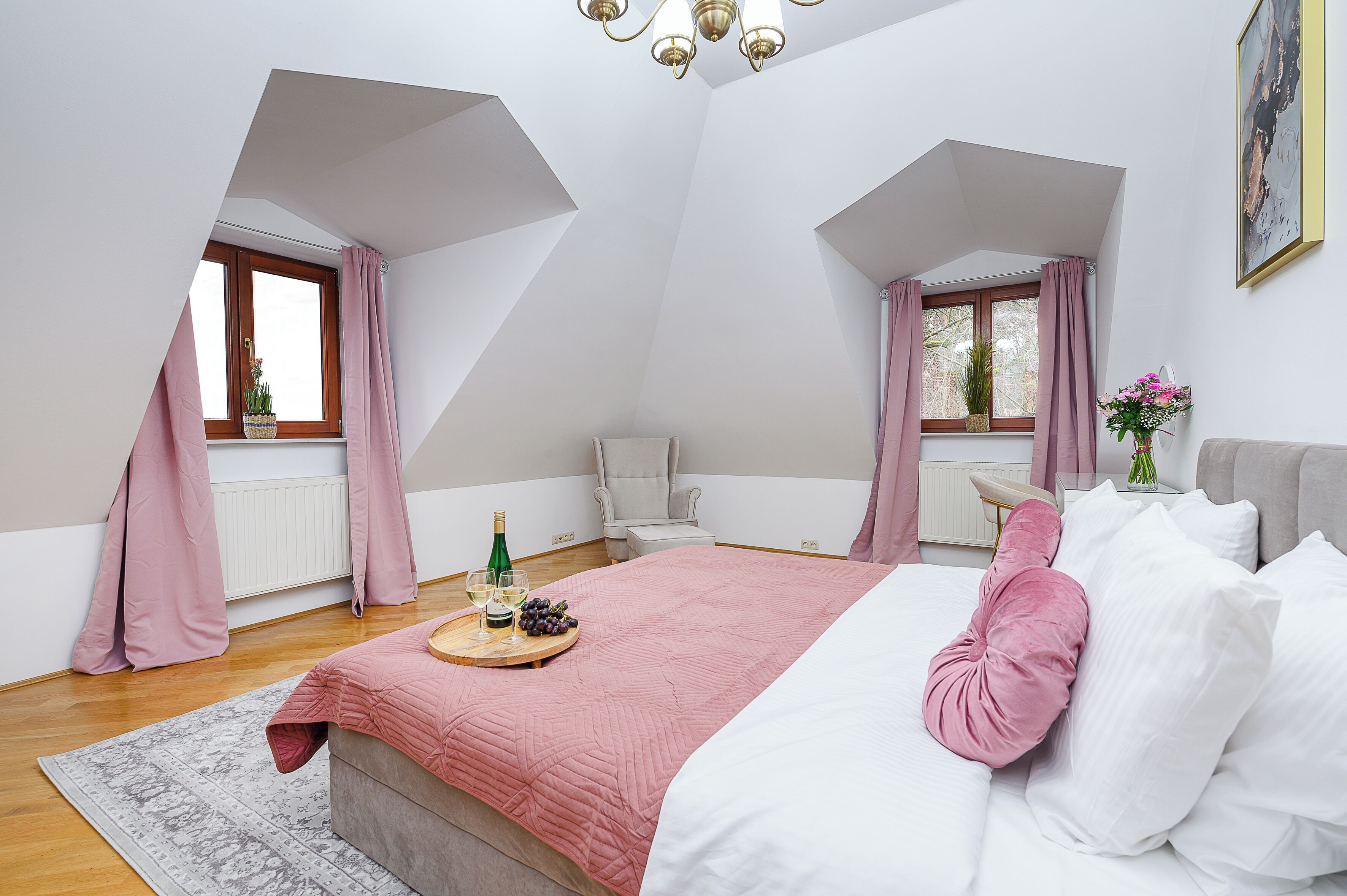 A bedroom with a sloped ceiling, a bed with a pink pillow, a bedside table with a bottle and glasses, and a window with curtains.