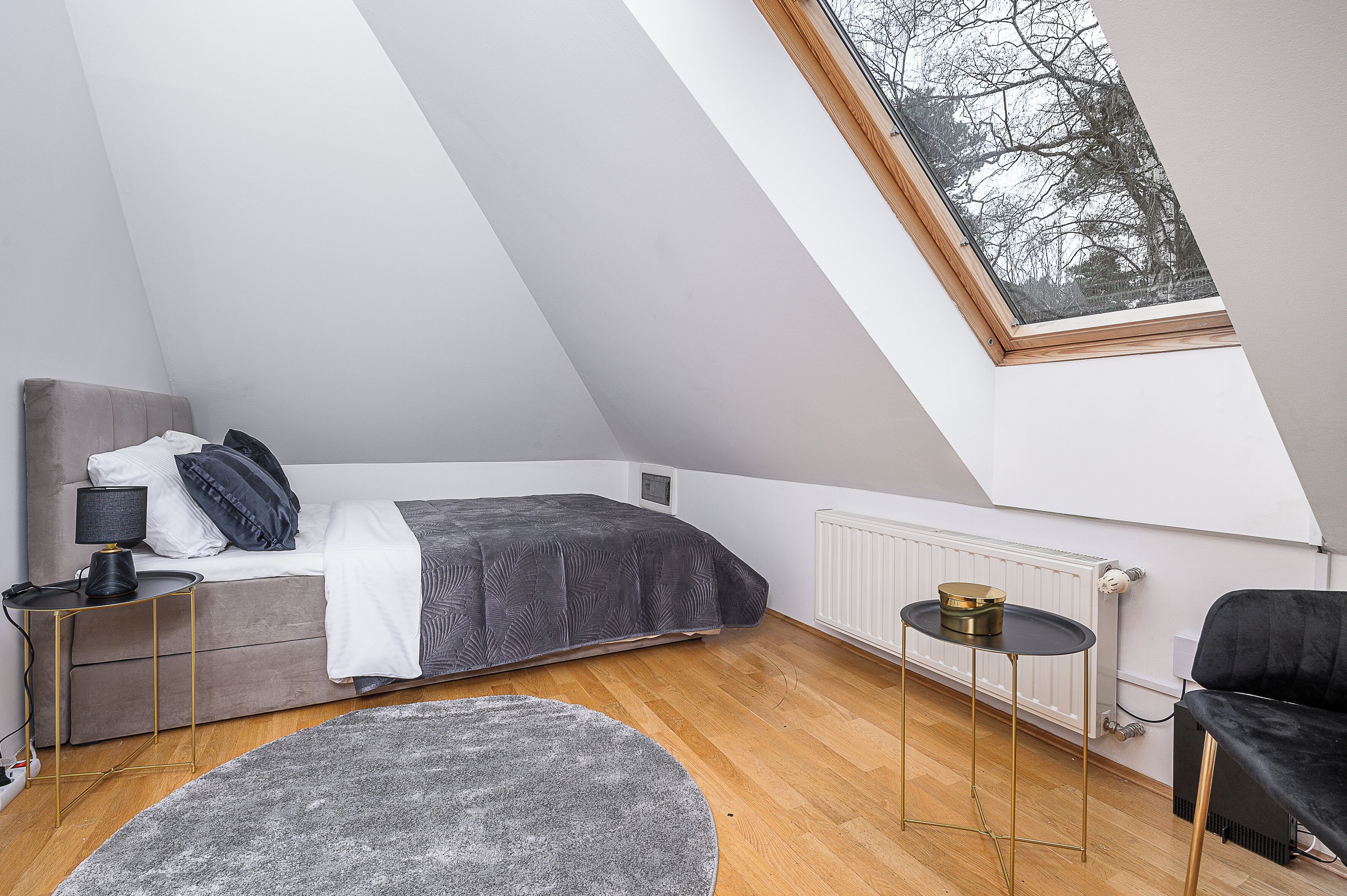 A bedroom with a sloped ceiling, a bed, a small table, a chair, a radiator, and a window with a view of trees.