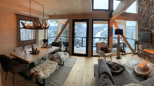 A cozy interior with a stone fireplace, a dining area with a wooden table and chairs, and a large window offering a view of snow-covered mountains. - Superior Chalet # 25 with Sauna (Turrach)