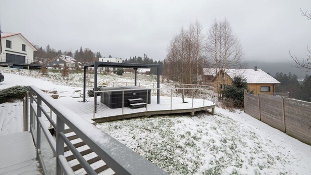 Un espacio exterior cubierto de nieve con una terraza de madera, barandilla metálica y un banco.