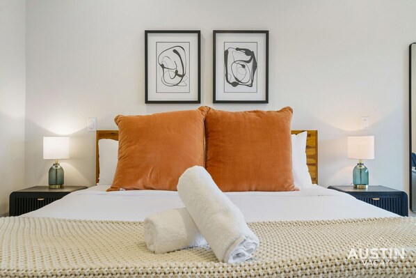 A neatly made bed with two pillows, a rolled towel, and two framed abstract artworks on the wall.