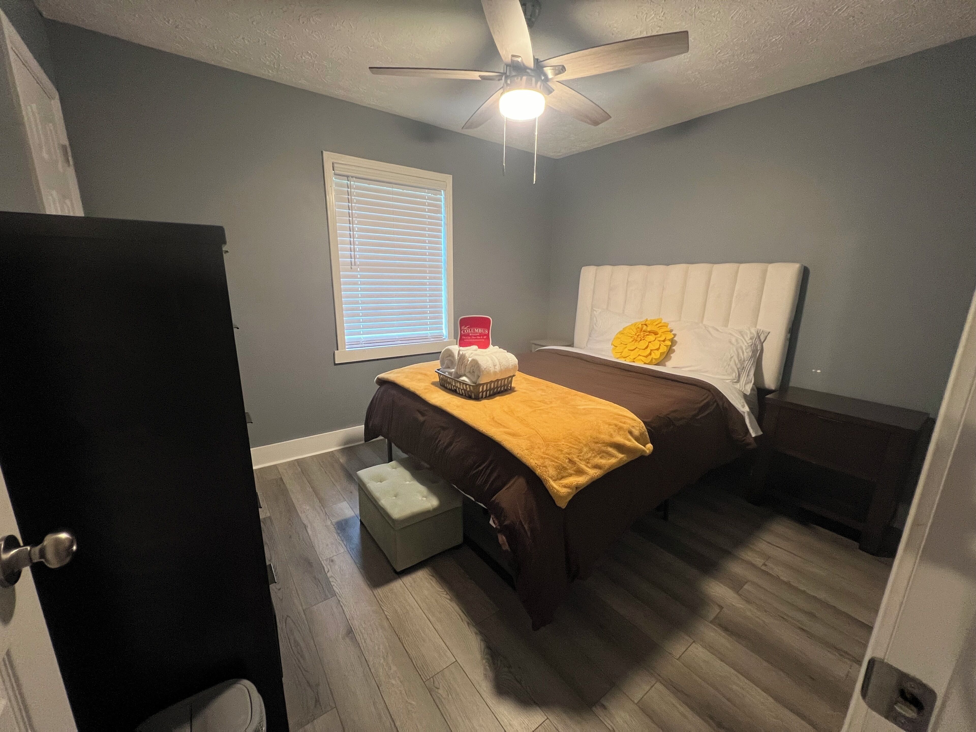 Une chambre à coucher comprenant un lit, une table de chevet, une fenêtre avec des stores, un ventilateur de plafond et une porte.