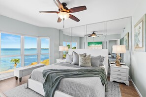 Une chambre à coucher avec un grand lit, deux tables de chevet, un ventilateur de plafond, une fenêtre donnant sur l’océan et un tableau accroché au mur.