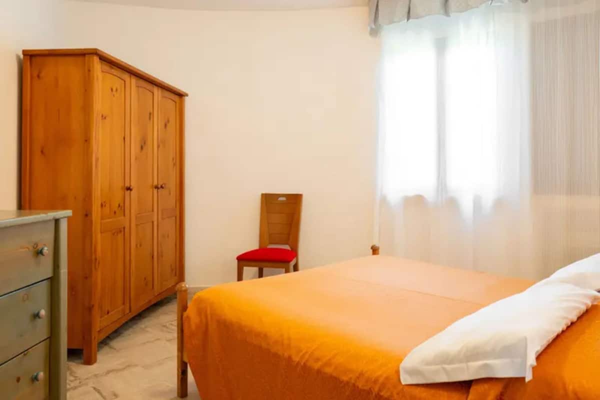A bedroom with a bed, a wooden wardrobe, a chair, and a window with curtains.