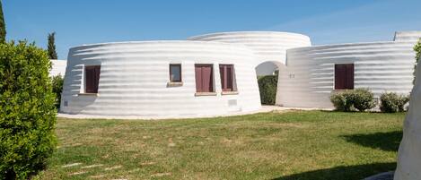 A cluster of white, dome-shaped buildings with small windows and shutters, surrounded by a grassy area with a clear sky.