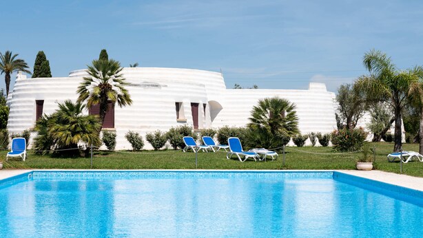 A swimming pool with lounge chairs, palm trees, and a white building with red shutters. - Hotiday Gallipoli Masseria (Tuglie)