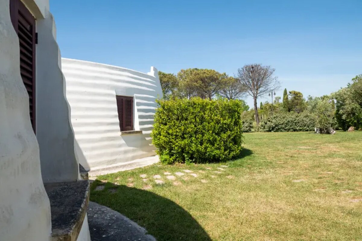 Un edificio bianco con una finestra e un portone, circondato da un prato curato e alberi.