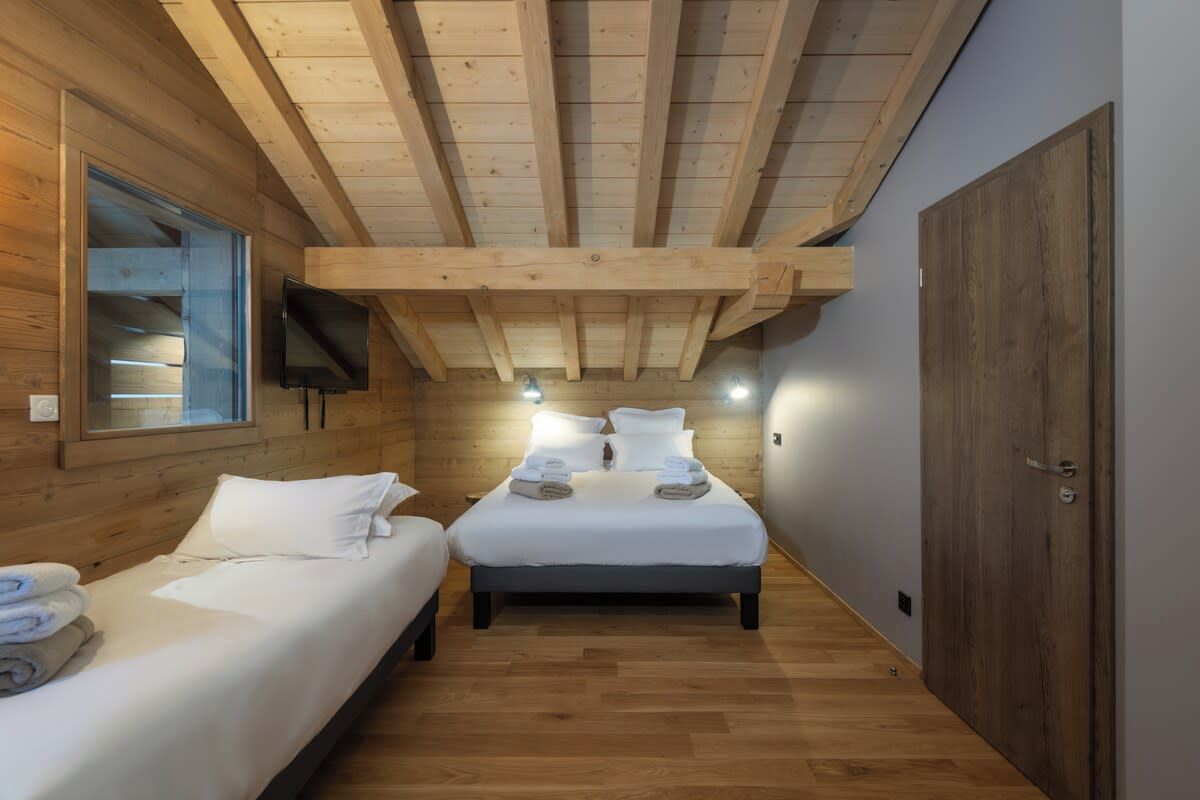 A room with two beds, wooden beams on the ceiling, a TV mounted on the wall, and a wooden door.