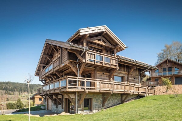 A multi-level wooden house with balconies and large windows, set on a grassy hillside.