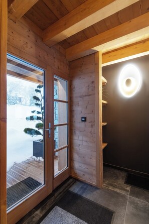 A wooden interior with a glass door leading outside, a dark wall with a circular light fixture, and a tiled floor.
