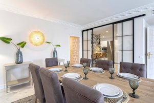 Comedor con una mesa de madera dispuesta para seis personas, rodeada de sillas tapizadas. Hay dos plantas grandes en macetas sobre la mesa y una lámpara colgante moderna ilumina el espacio.