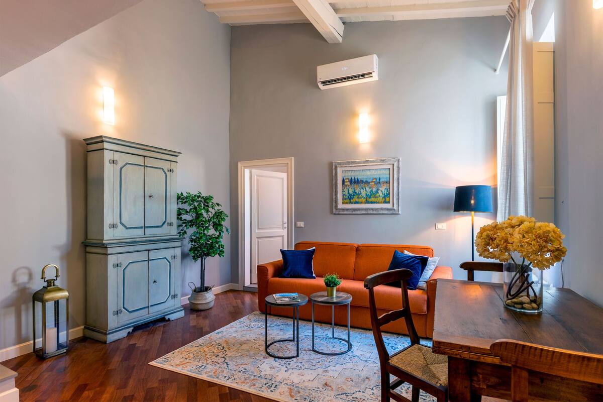 Una sala de estar con un sofá, dos mesitas auxiliares, un aparador, una planta en maceta y un jarrón con flores.