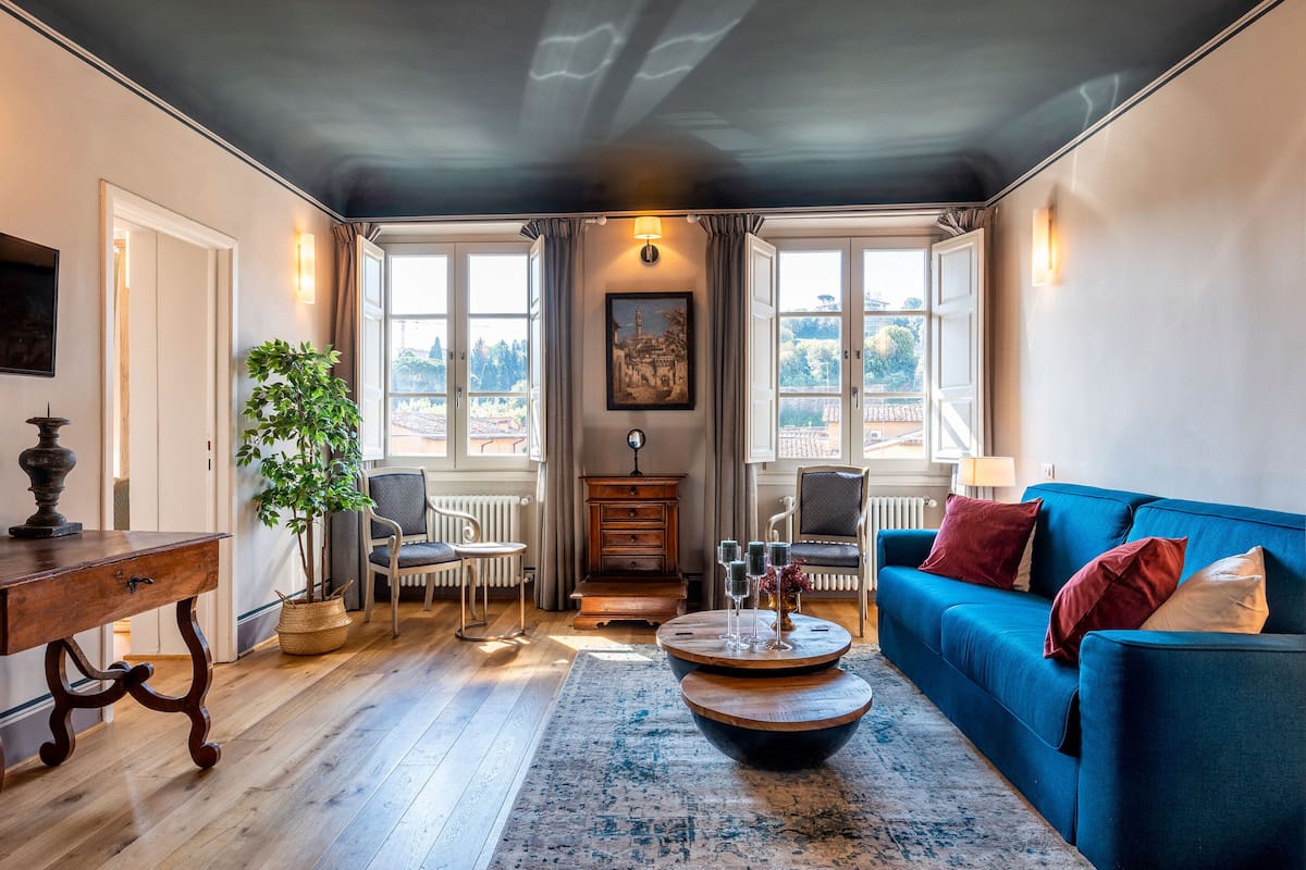 Una sala de estar acogedora con un sofá azul, una mesa de centro de madera, una planta y vistas al exterior a través de amplios ventanales.