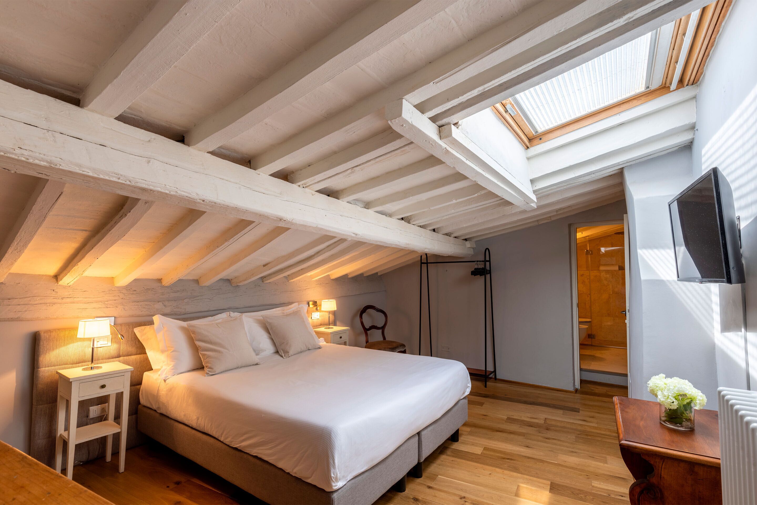 Un dormitorio con cama, mesitas de noche, lámpara, sillón, televisor y tragaluz.