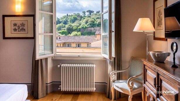 A hotel room with a large window, a radiator, a chair, a desk with a lamp, and a framed picture on the wall. - Genteel Home Piazza Pitti (Florence)