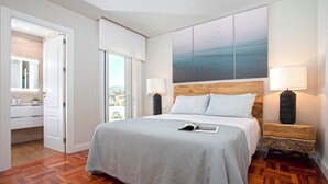 Un dormitorio con una cama grande, dos mesitas de noche con lámparas, vistas al mar a través de una ventana y suelo de madera.