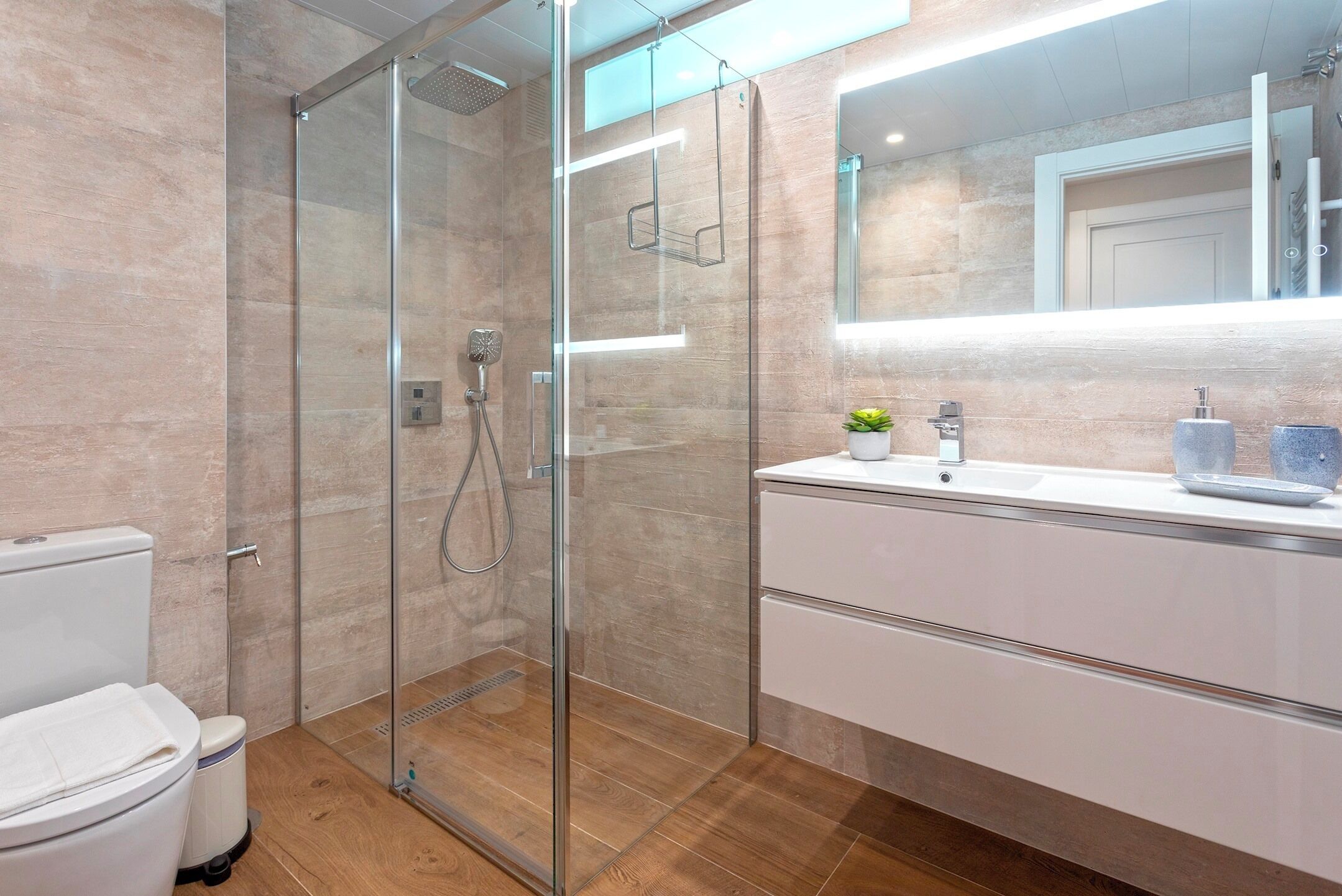 Een moderne badkamer met een glazen douchecabine, een witte wastafel met spiegel erboven en een houten vloer.