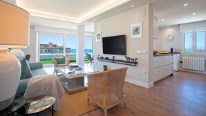 Una sala moderna con sofá, mesa de centro, silla de mimbre, televisor montado en la pared y una cocina con mostrador y ventana.