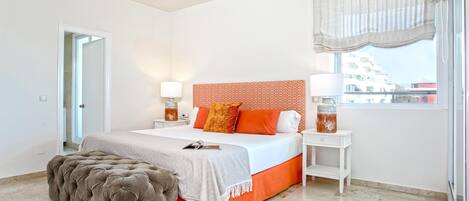 A bedroom with a large bed, a nightstand, a lamp, a window with curtains, and a view of buildings outside.