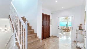 Un escalier avec des balustrades blanches menant à une porte en bois, un coin repas offrant une vue sur la mer, et une console blanche agrémentée d’un vase et de bougies.