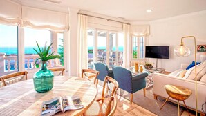 Un salon moderne doté d’une grande fenêtre donnant sur la mer, un coin repas avec une table et des chaises, et un canapé avec un téléviseur.