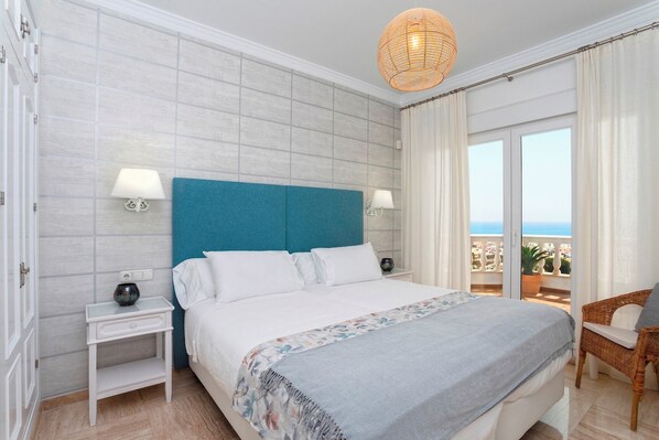 A bedroom with a large bed, two bedside tables, a nightstand, a wicker chair, and a view of the sea through a balcony.