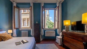 A bedroom with a bed, a dresser, a television, and two windows offering views of a building and trees. - Genteel Home Biblioteca Nazionale (Florence)