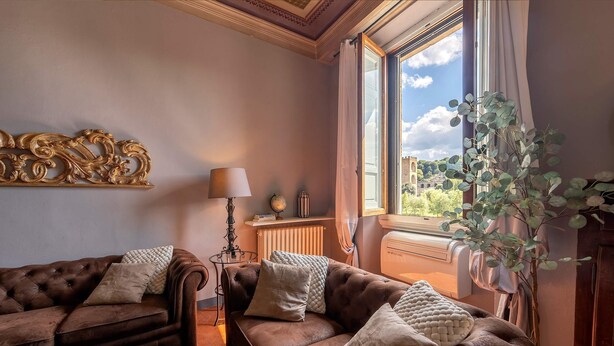 A cozy living room with two brown sofas, a small table, a lamp, and a window with a view of greenery and a distant building. - Genteel Home Biblioteca Nazionale (Florence)