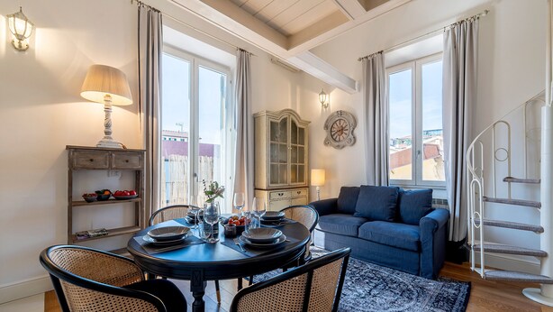 A dining area with a round table set for four, surrounded by wicker chairs, a cabinet with decorative items, and a blue sofa in the background. - Genteel Home Scarlatti (Florence)