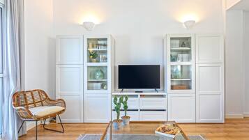 Un salon moderne avec un téléviseur, des meubles blancs, un fauteuil en osier et une table basse.