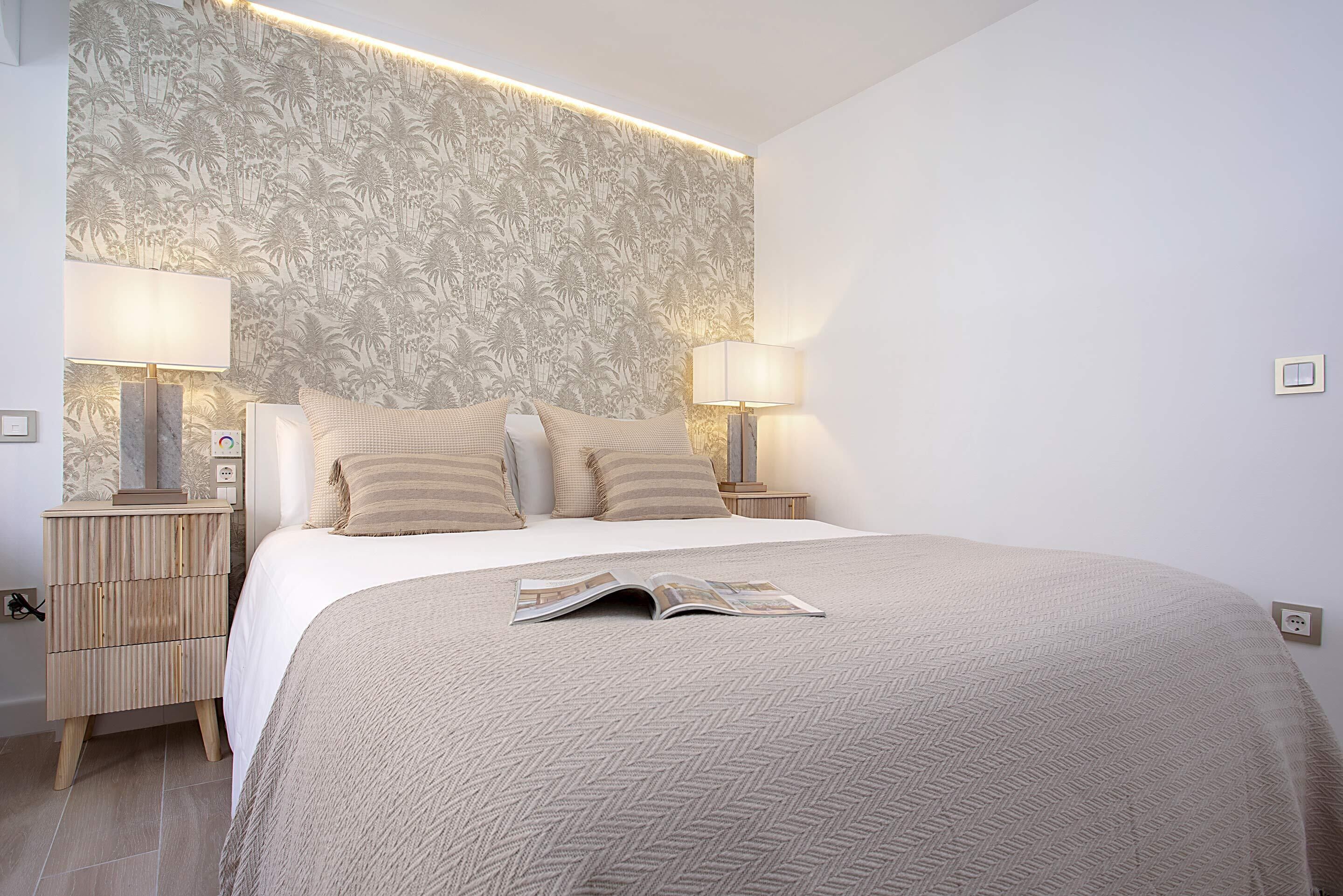 Un dormitorio con una cama, dos mesitas de noche, una mesilla auxiliar, una pared con estampado floral y una revista sobre la cama.