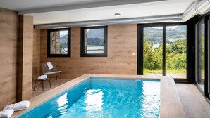Una piscina interior moderna con paredes de madera y grandes ventanales que ofrecen una vista panorámica de colinas y un lago.