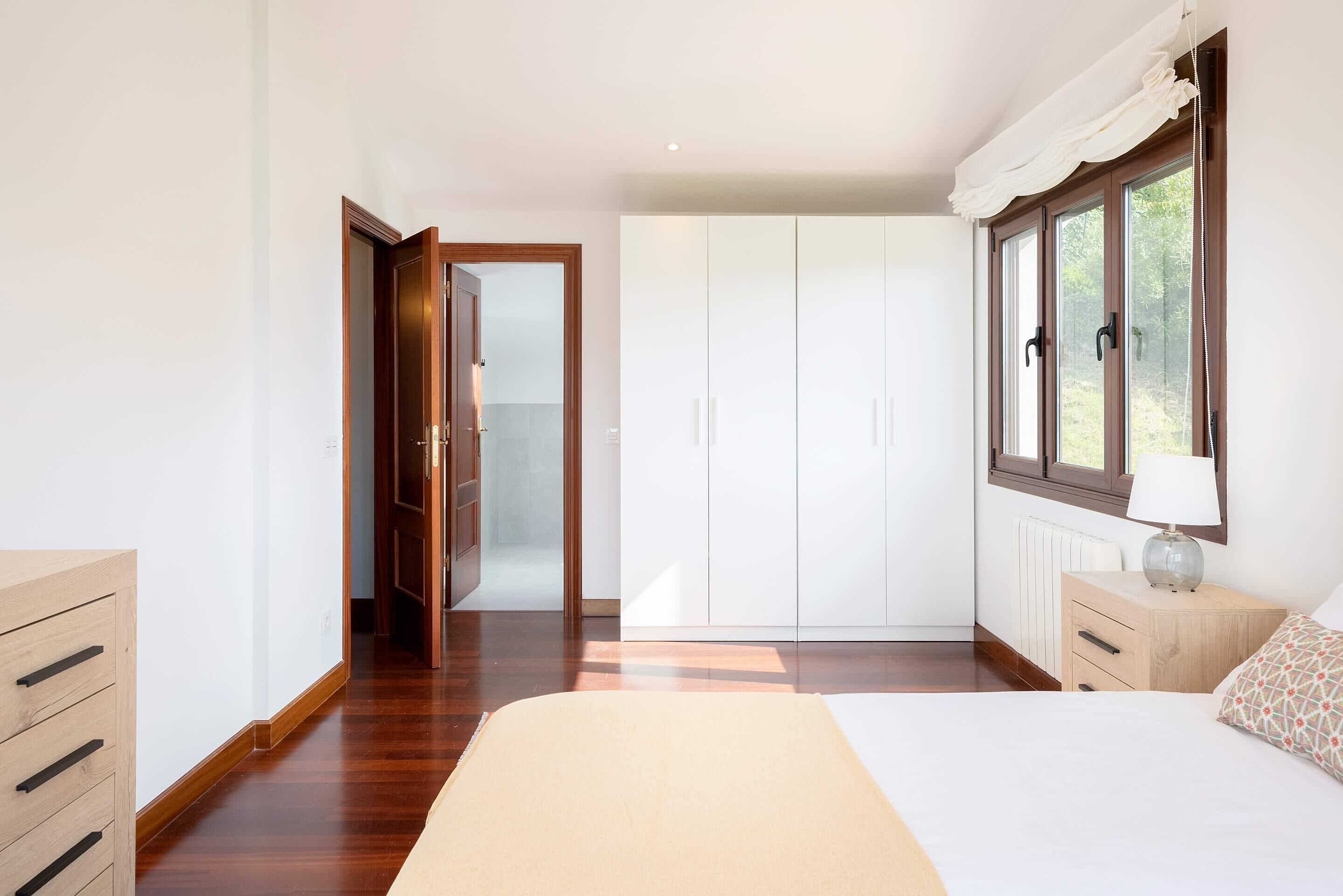 A bedroom with a bed, a wardrobe, a bedside table with a lamp, and a window with curtains.