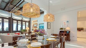 A dining area with a glass table set for six, surrounded by wooden chairs. There are wicker pendant lights above the table, and a view of the living room with a sofa and a dining table in the background. - Genteel Home Paseo Marítimo (Málaga)