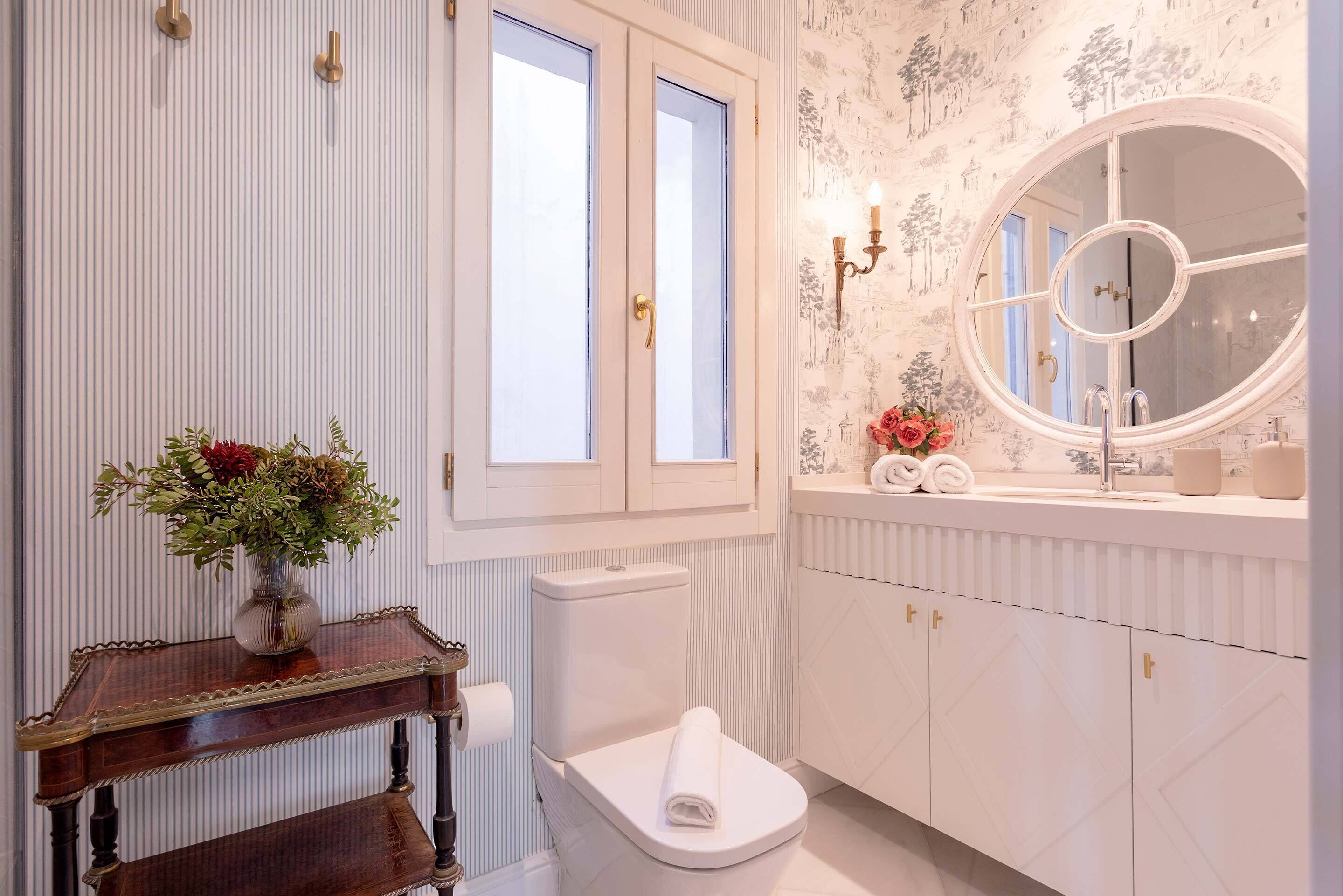 A bathroom with a toilet, a vanity with a mirror, a vase with flowers, and a window.
