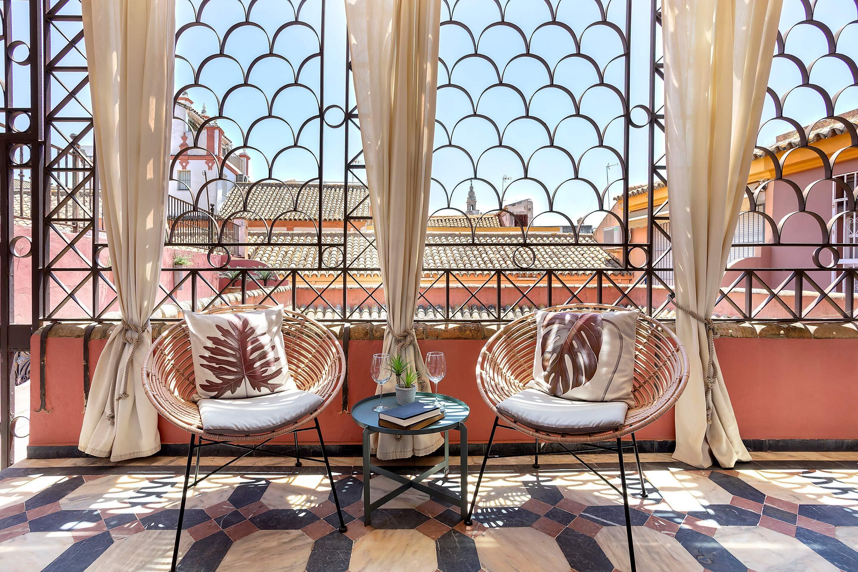 A terrace with two wicker chairs, a small table, and a view through a metal gate with a tiled roof in the background.