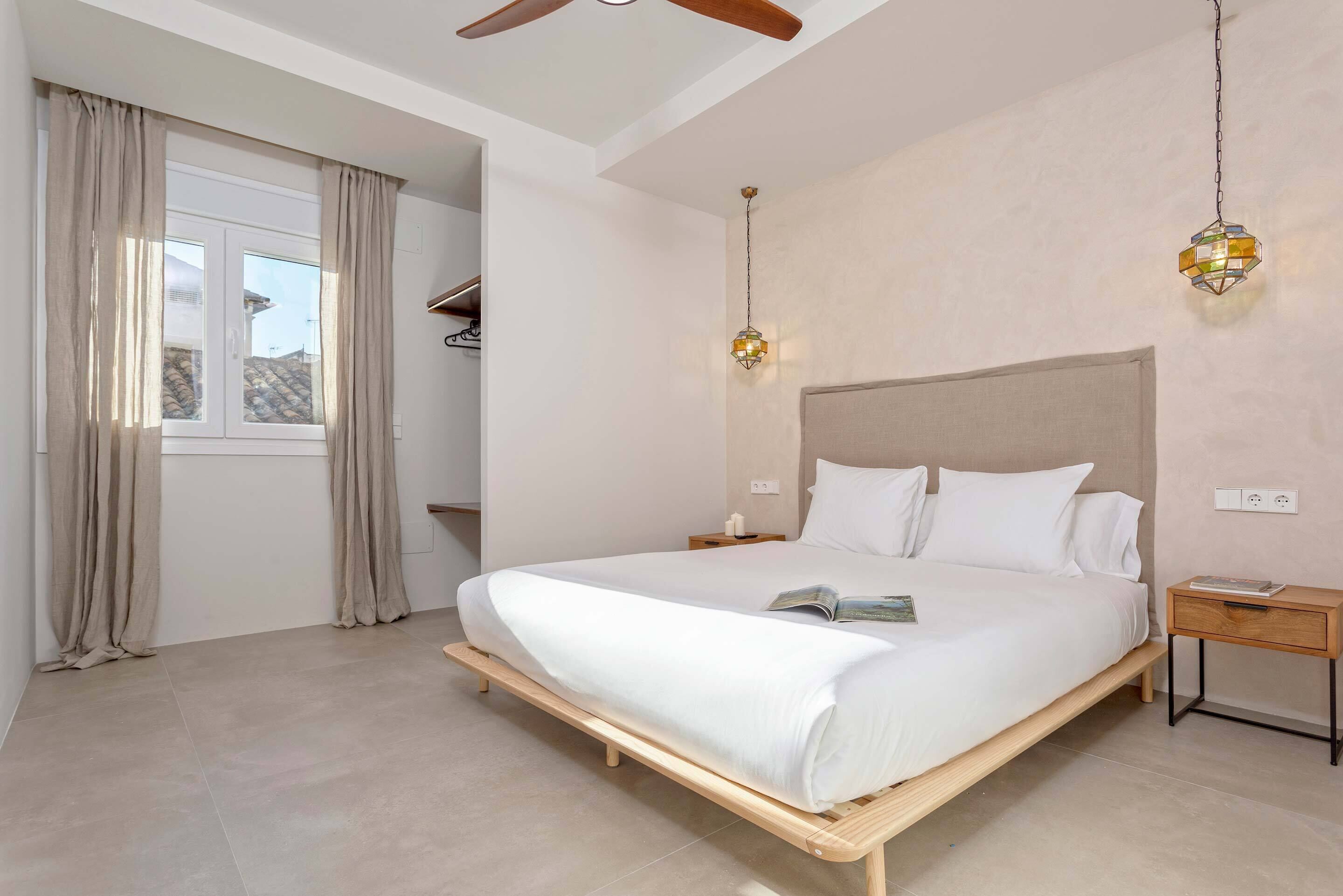 A bedroom with a bed, a nightstand, a bedside table, a ceiling fan, a window with curtains, and a hanging light fixture.