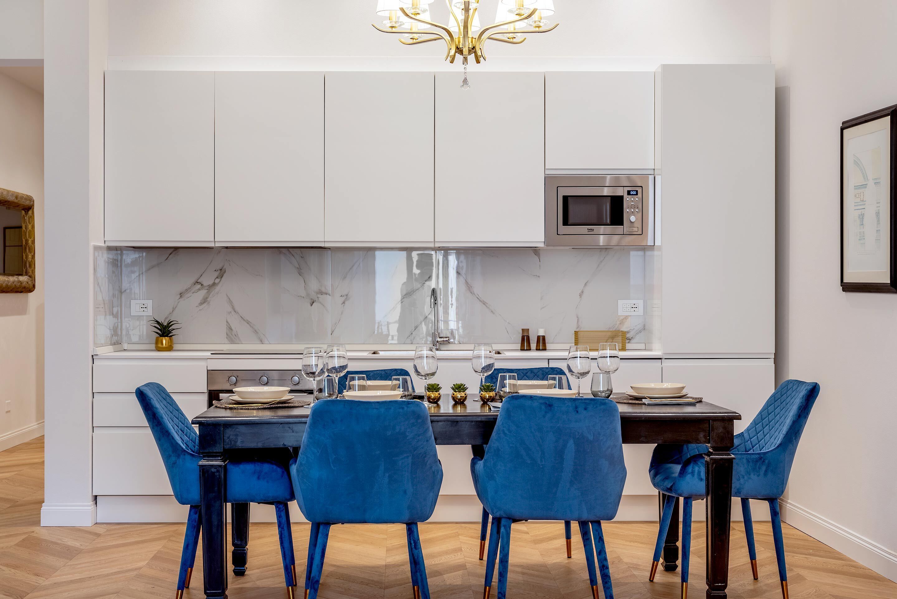 Cozinha moderna com armários brancos, micro-ondas embutido e painel de parede em mármore. Há uma mesa de jantar com cadeiras azuis e um lustre acima.