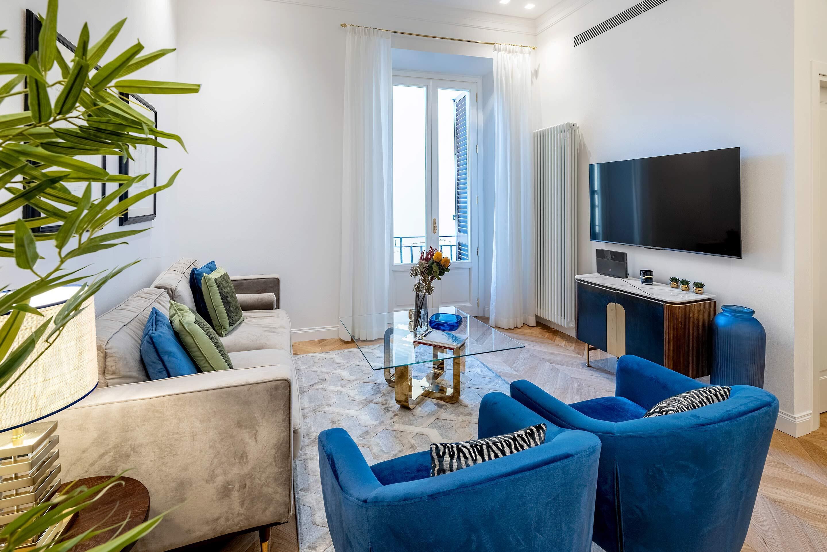 Uma sala de estar moderna com um sofá, duas poltronas azuis, uma mesa de centro de vidro, uma televisão e uma pequena mesa lateral.