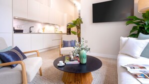 A modern living room with a round coffee table, a sofa, and a chair. There is a kitchen in the background with white cabinets and a large flat-screen TV mounted on the wall. - Genteel Home Plaza del Obispo (Málaga)