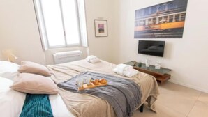Ein ordentlich eingerichtetes Hotelzimmer mit Bett, Nachttisch mit Frühstücksangebot und einem Flachbildfernseher. Ein Fenster mit Jalousien ist ebenfalls zu sehen.