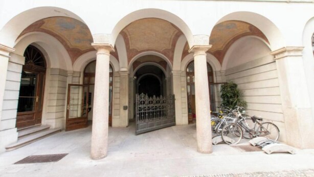 Ein Gebäude mit einem zentralen Eingang, der von drei Bögen gesäumt wird, einem befestigten Bereich und Fahrrädern, die davor abgestellt sind.
