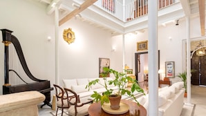Een ruime woonkamer met een vleugelpiano, een witte bank, een ronde houten tafel met een plant en een trap naar een hoger gelegen verdieping.