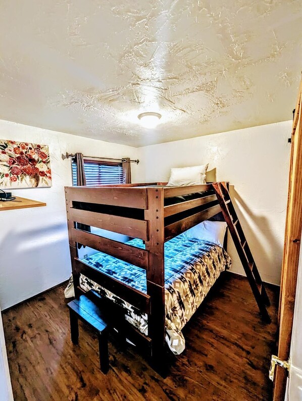 Una cama individual sobre otra en una habitación con suelo de madera, luz de techo, ventana con persianas y estante adosado a la pared.