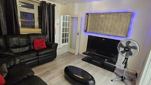 A living room with a black leather sofa, a red pillow, a flat-screen TV, and a standing fan. - Spacious & Modern 2-bed House in Dagenham (Dagenham)